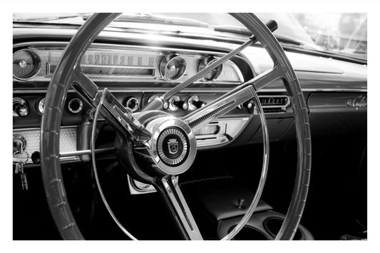 Black and white close-up photograph of a vintage car interior, featuring a large three-spoke steering wheel with chrome trim and a dashboard with round gauges.