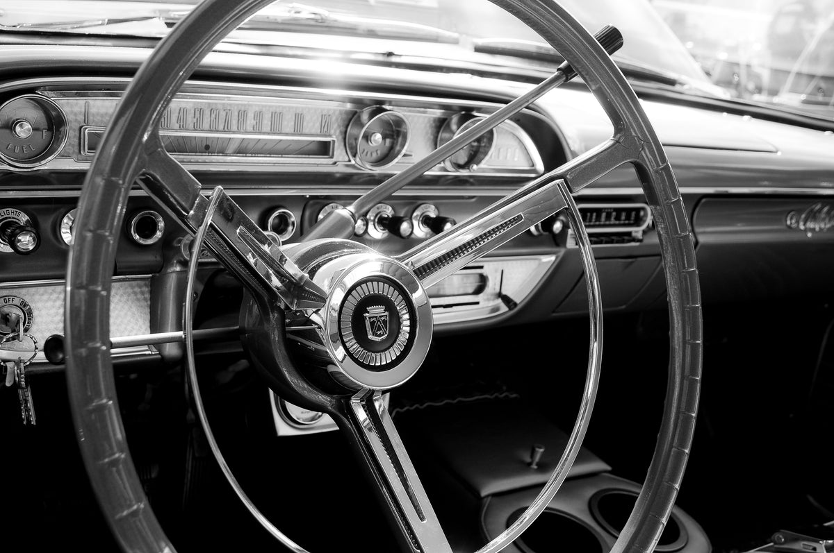 Black and white close-up of a vintage car interior featuring a large three-spoke steering wheel and dashboard instruments.