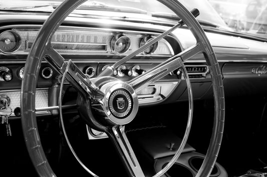 Black and white close-up of a vintage car interior featuring a large three-spoke steering wheel and dashboard instruments.